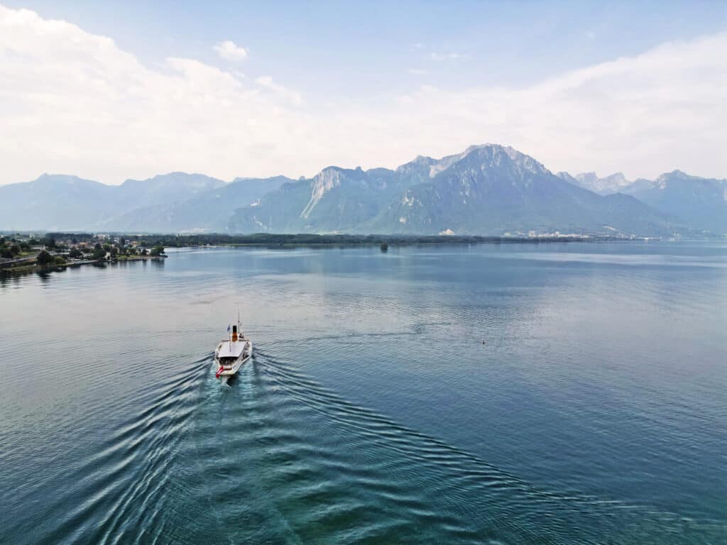 Vue aérienne d'un bateau à vapeur Belle Époque de la CGN naviguant sur les eaux calmes du lac Léman, laissant un sillage en V, avec les Alpes suisses et leurs sommets majestueux en arrière-plan sous un ciel partiellement nuageux