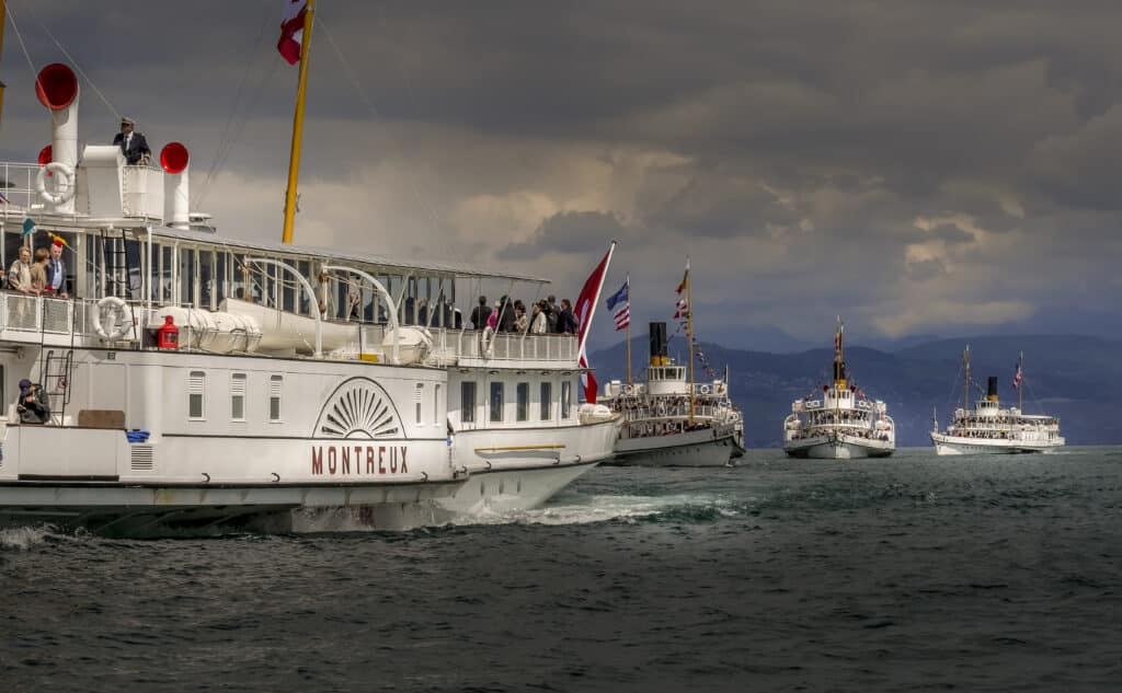 Le bateau à vapeur Montreux de la CGN en premier plan avec son nom visible sur la coque blanche et son logo en éventail, passagers sur les ponts, suivi de trois autres bateaux Belle Époque en formation, pavillons suisses déployés, ciel orageux dramatique et montagnes sombres en arrière-plan sur le lac Léman