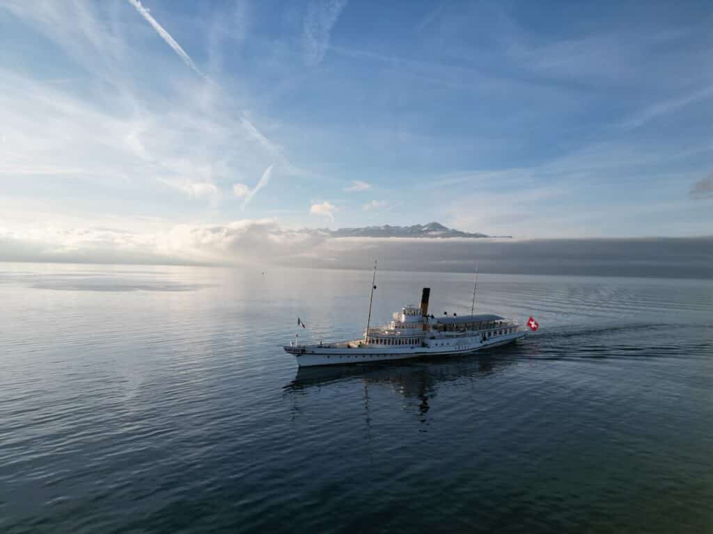 Bateau à vapeur Belle Époque Vevey de la CGN avec pavillon suisse naviguant sur les eaux calmes du lac Léman au lever du soleil, brume matinale sur les montagnes en arrière-plan, ciel bleu parsemé de traînées d'avion et lumière dorée rasante