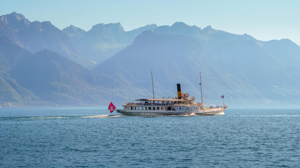 Le bateau à vapeur Belle Époque Vevey de la CGN naviguant de profil sur les eaux calmes du lac Léman, coque blanche élancée, cheminée dorée, pavillon suisse à la poupe, passagers visibles sur les ponts, imposante chaîne des Alpes brumeuses en arrière-plan, atmosphère paisible