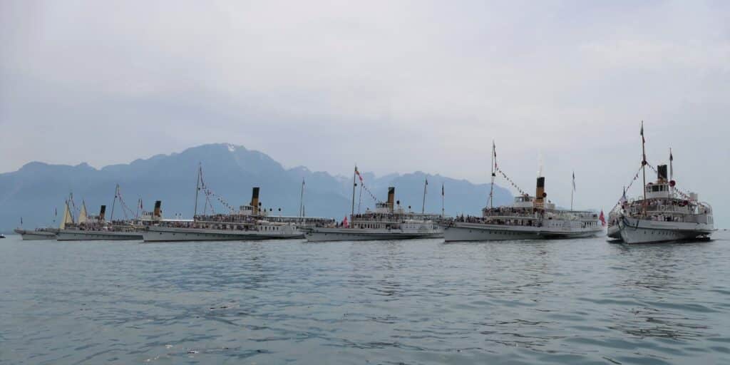 Six bateaux à vapeur Belle Époque de la CGN naviguant en ligne sur les eaux bleues du lac Léman sous un ciel ensoleillé avec nuages blancs, coques blanches, cheminées sombres, pavillons déployés, montagnes lointaines à l'horizon