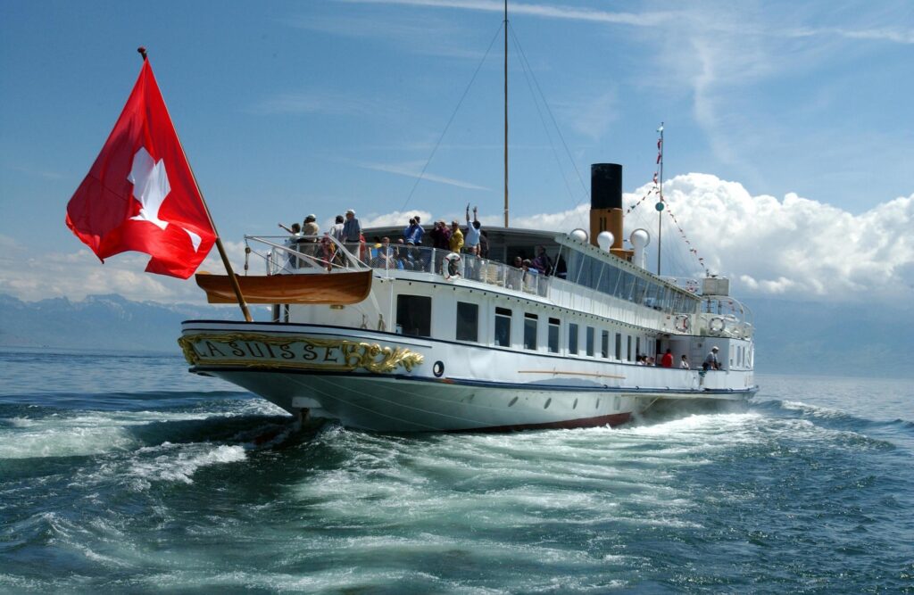 Vue arrière (poupe) du bateau à roues à aubes La Suisse naviguant sur le Lac Léman. Le nom "LA SUISSE" est inscrit en lettres dorées entourées de sculptures ornementales. Un grand drapeau suisse flotte à l'arrière, et de nombreux passagers profitent du pont supérieur.