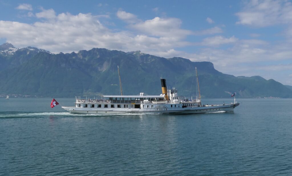Vue de côté du bateau à roues à aubes Italie de la CGN en pleine navigation sur le Lac Léman par temps ensoleillé. Le navire blanc et jaune, l'un des plus célèbres de la flotte Belle Époque, a pour arrière-plan les majestueuses montagnes de la Riviera.