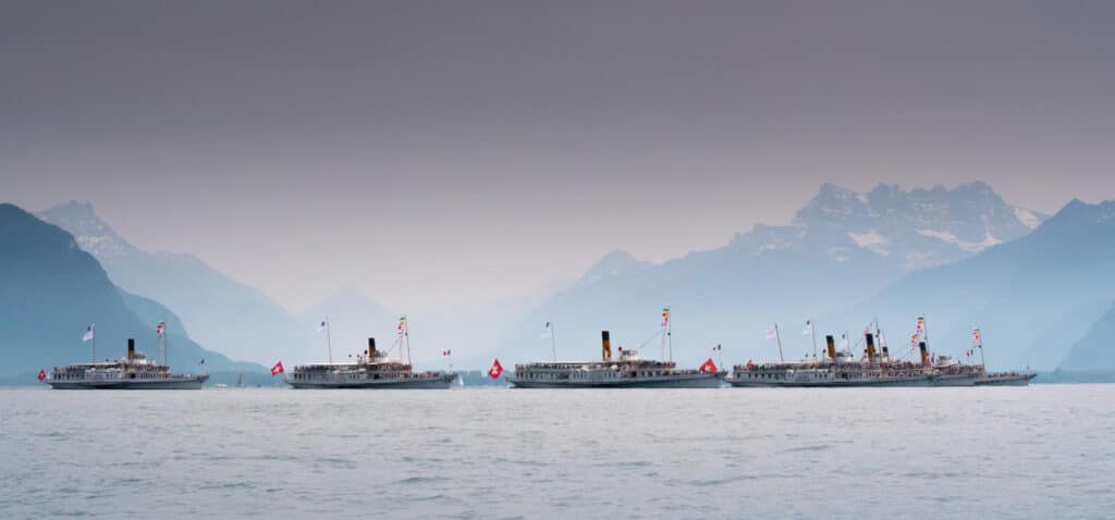 Six bateaux à vapeur Belle Époque de la CGN en formation sur le lac Léman au crépuscule, coques blanches, cheminées fumantes, pavillons suisses, passagers à bord, sommets alpins enneigés des Dents du Midi visibles dans la brume rosée du soir