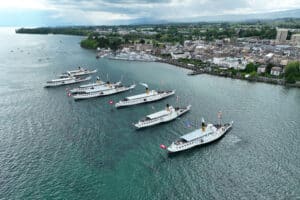 Vue aérienne des six bateaux à vapeur Belle Époque de la CGN naviguant en formation serrée devant les quais de Morges, sur le Lac Léman. Célébration de la flotte des bateaux à roues à aubes classés monuments historiques.